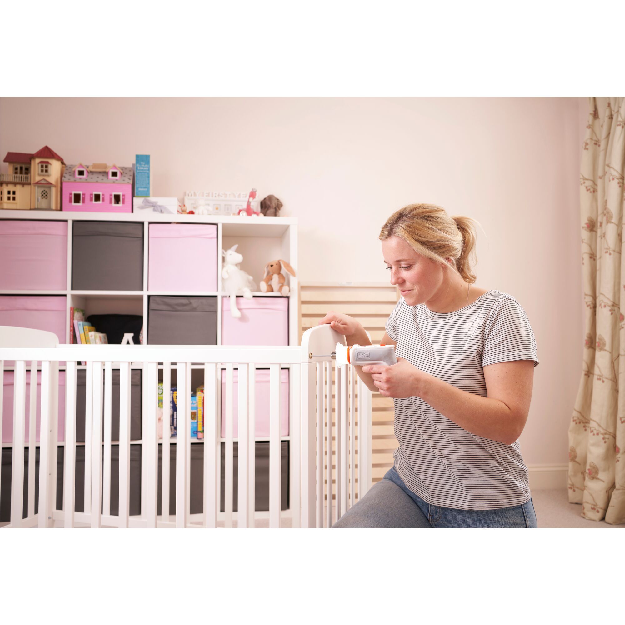A person is using a BLACK+DECKER tool to assemble or adjust a white baby crib in a nursery. The background features a storage unit with pink and grey boxes, toys, books, and a dollhouse. The person's face is obscured for privacy.