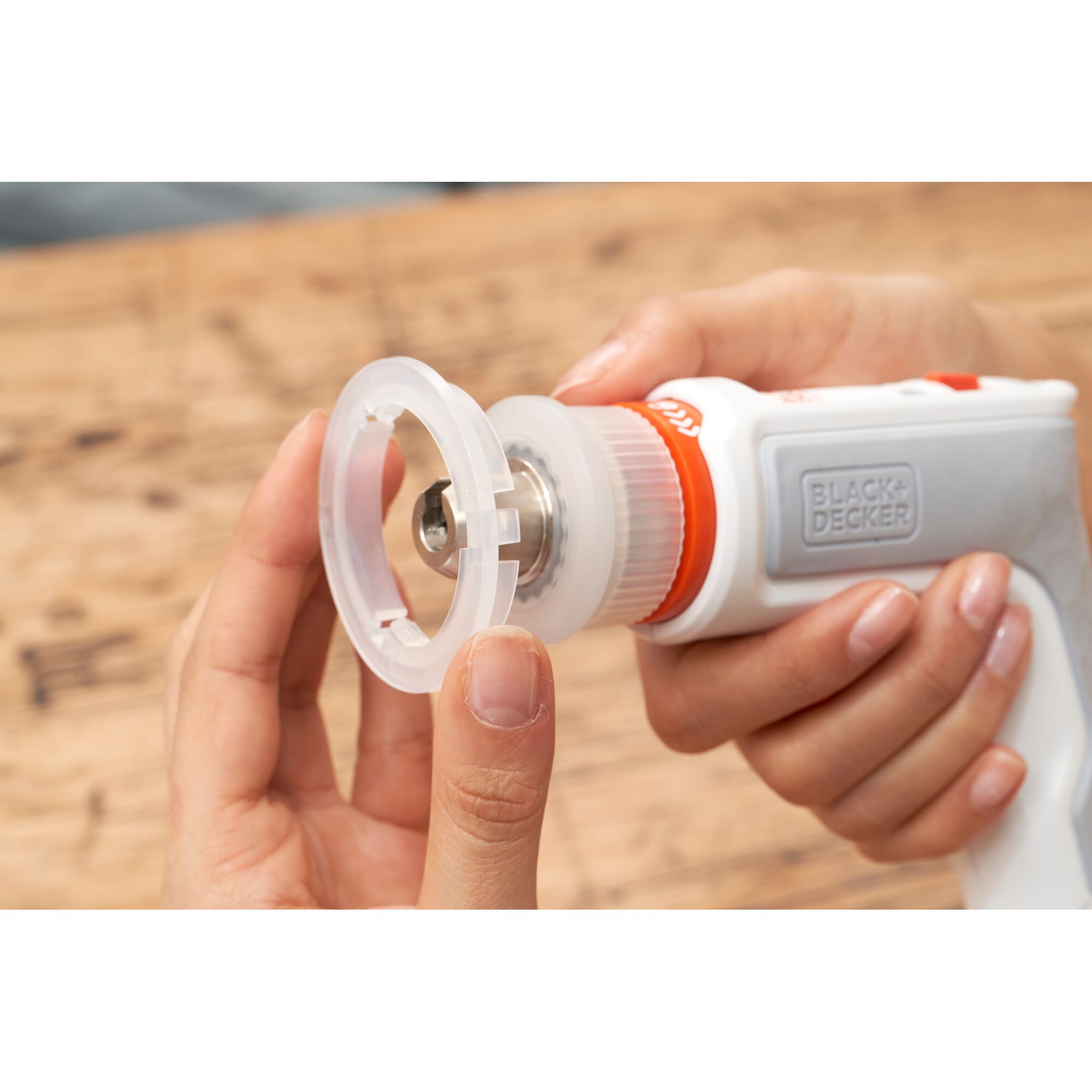 Close-up of a person assembling an attachment onto a BLACK+DECKER white handheld tool, likely part of a photography-related equipment, on a wooden surface.
