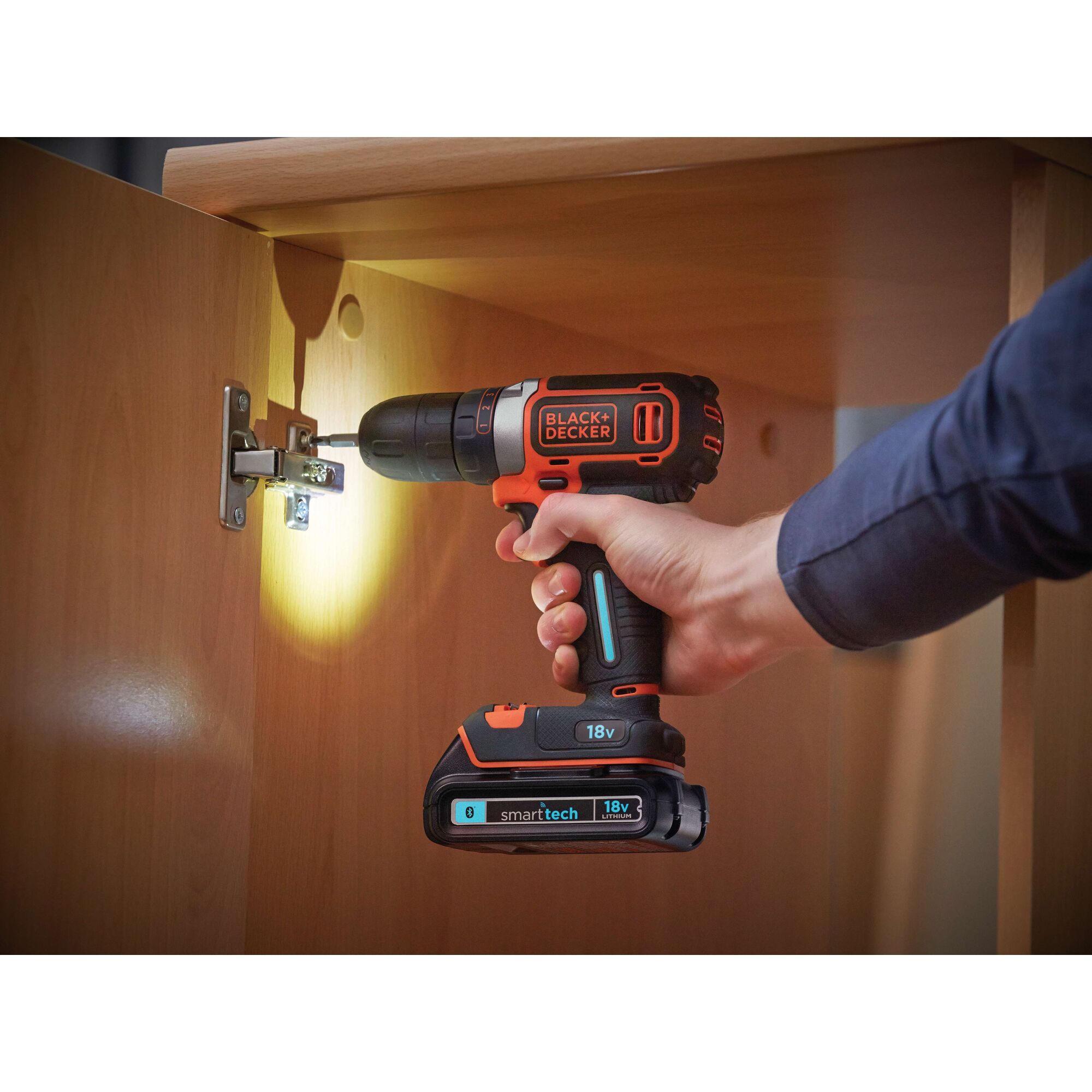 A person using a BLACK+DECKER 18V cordless drill to install a hinge on a wooden cabinet. The drill is equipped with a 'smartech' battery and an LED light illuminating the working area.