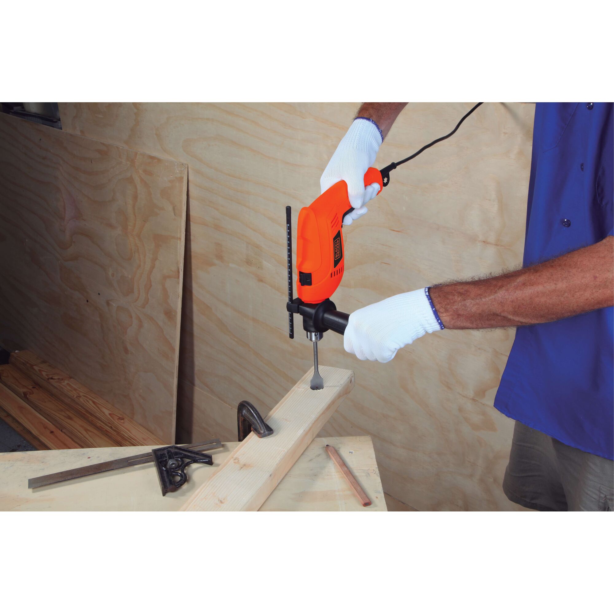 A person wearing white gloves is using a BLACK+DECKER HD555 orange electric drill to make a hole in a piece of wood in a workshop setting. Measuring tools and a clamp are placed nearby.