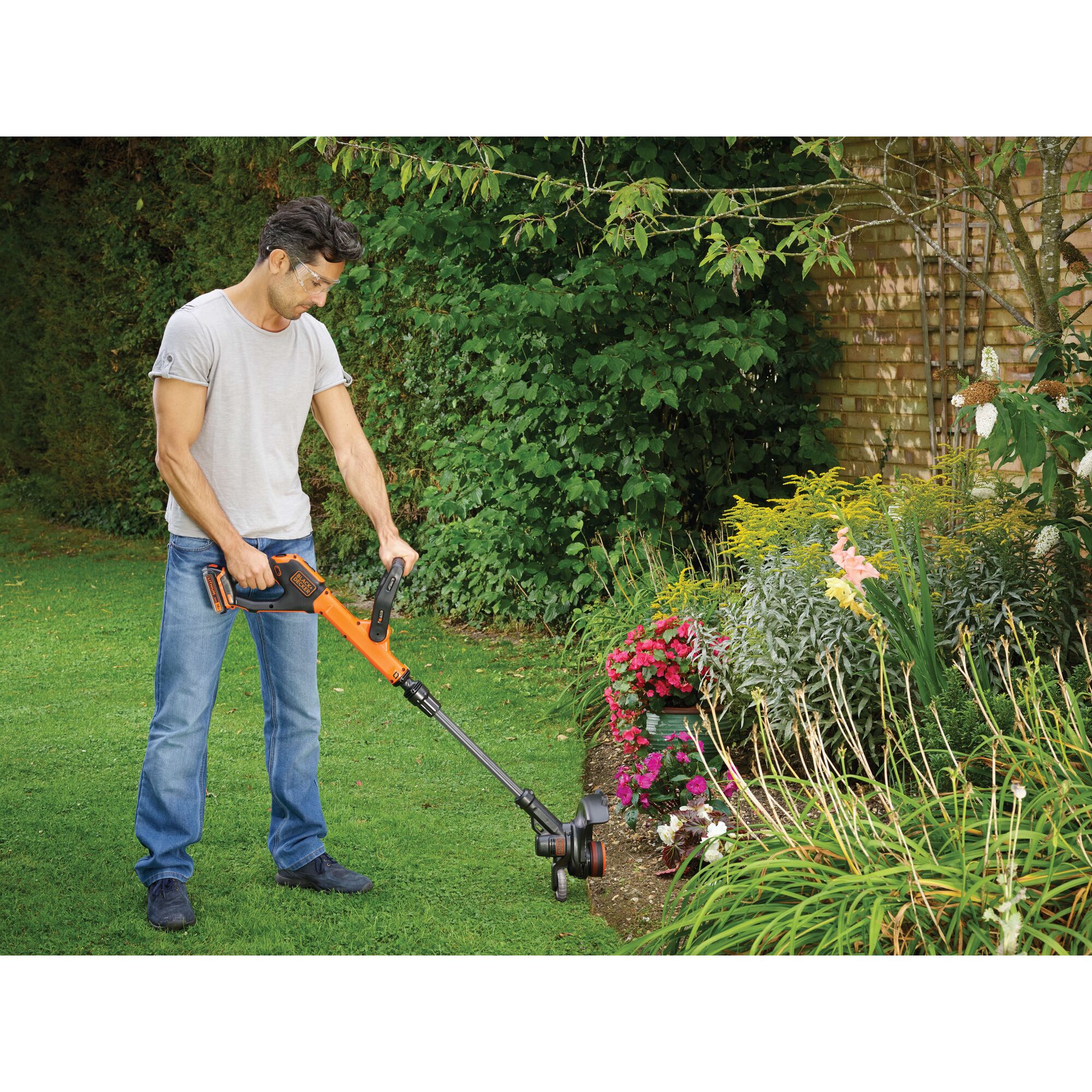 A person is using a BLACK+DECKER cordless orange grass trimmer in a garden, trimming the edge of a flower bed. The person's face is blurred for privacy. The garden is lush and features various flowering plants and greenery.