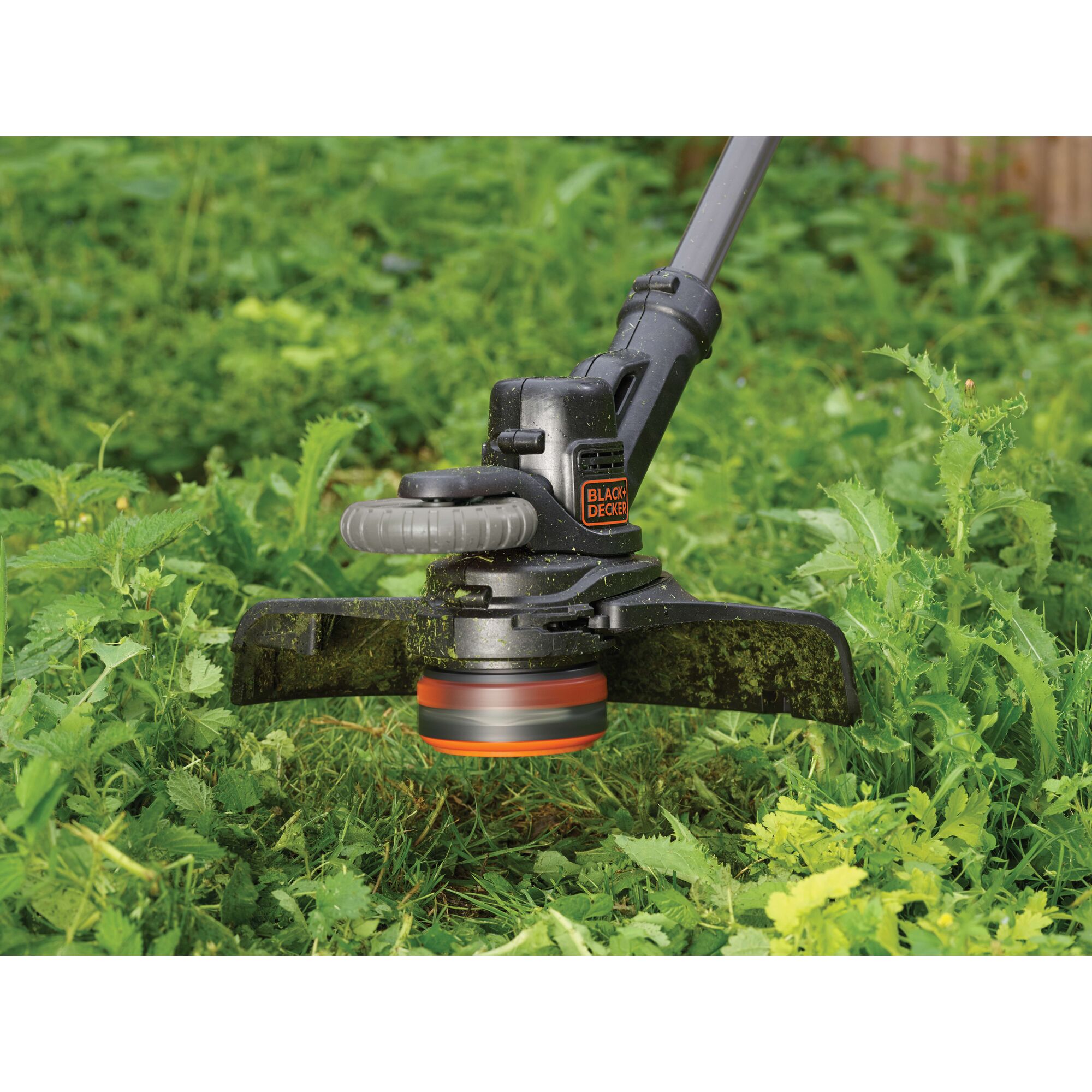 Close-up of a BLACK+DECKER cordless grass trimmer in use, cutting green grass and weeds outdoors.