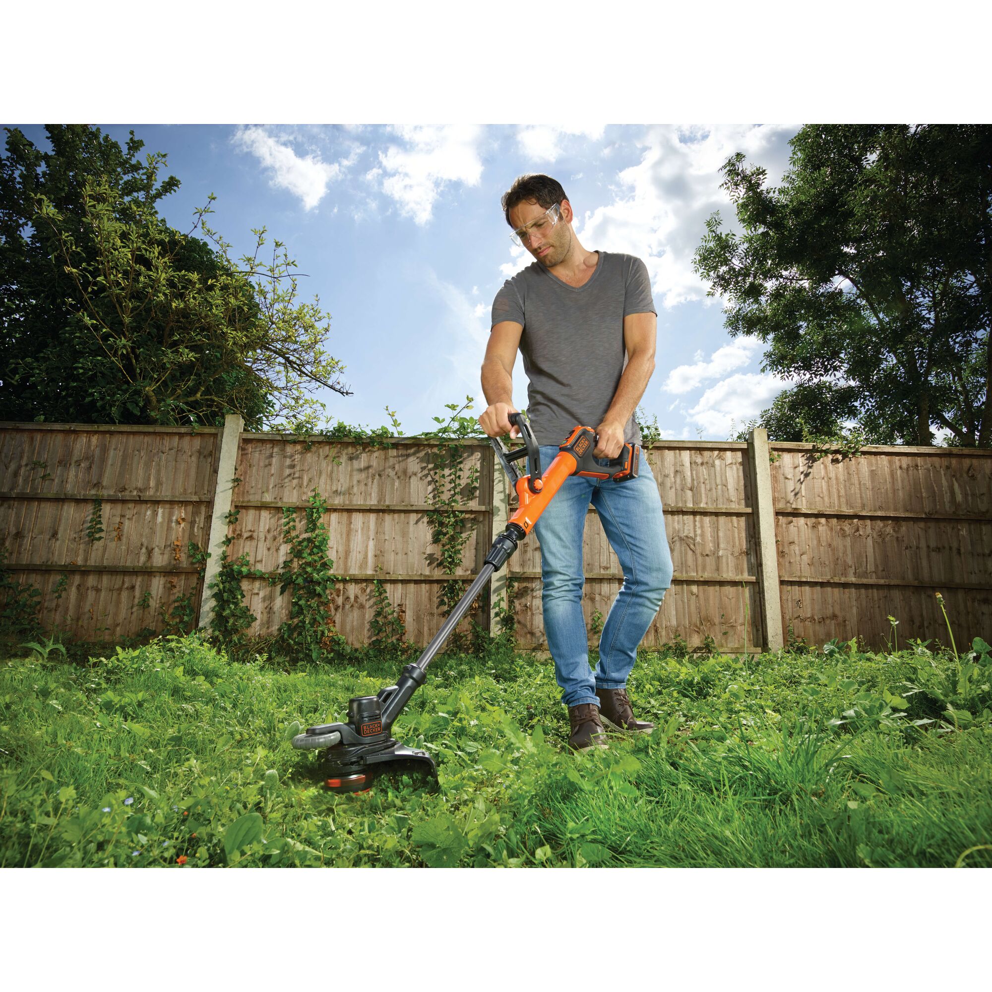 Person using a BLACK+DECKER cordless grass trimmer (model STC1820EPCF) in a garden with overgrown grass and a wooden fence in the background.