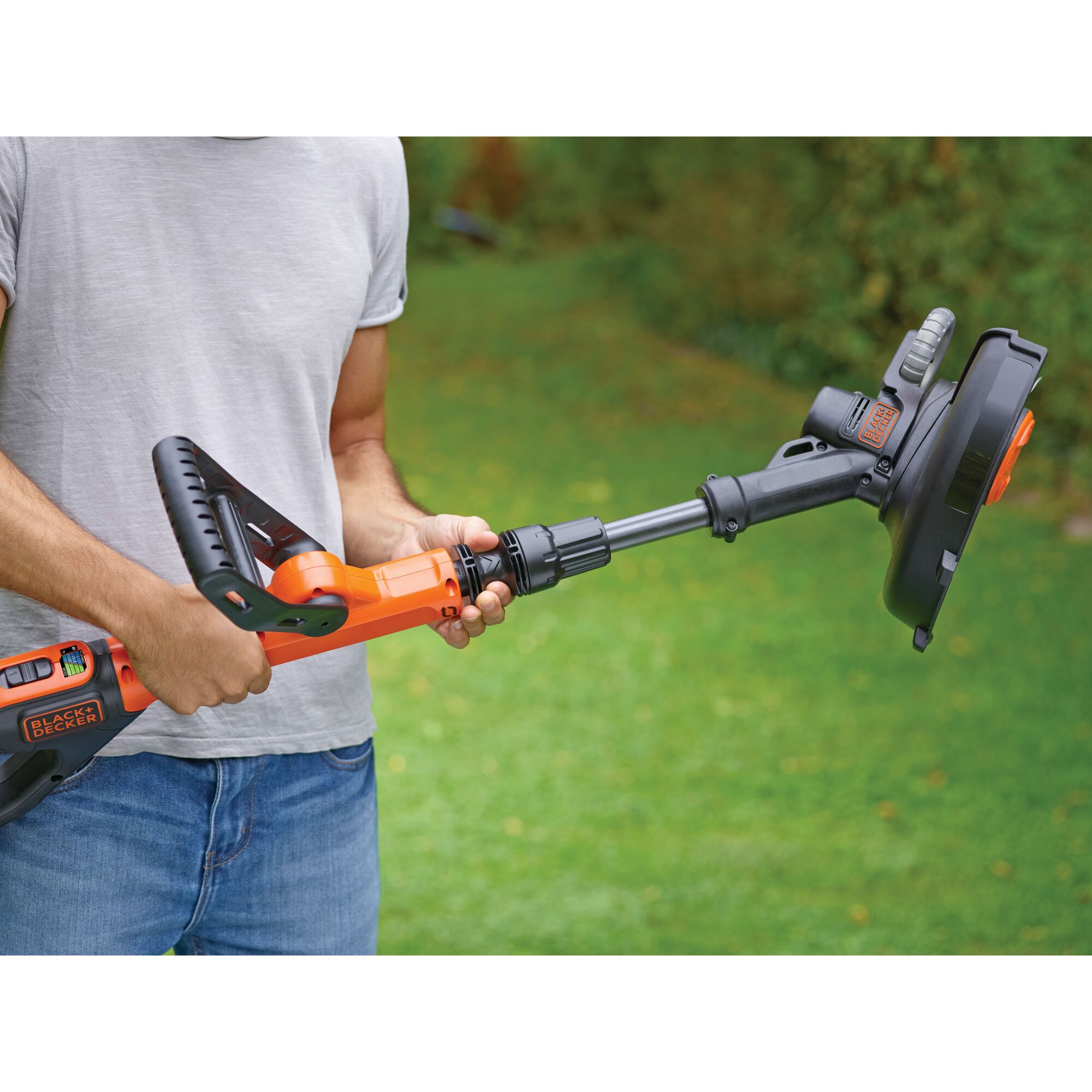 A person is holding a BLACK+DECKER cordless grass trimmer in a garden. The trimmer is orange and black, and the person is wearing a light grey t-shirt and blue jeans.